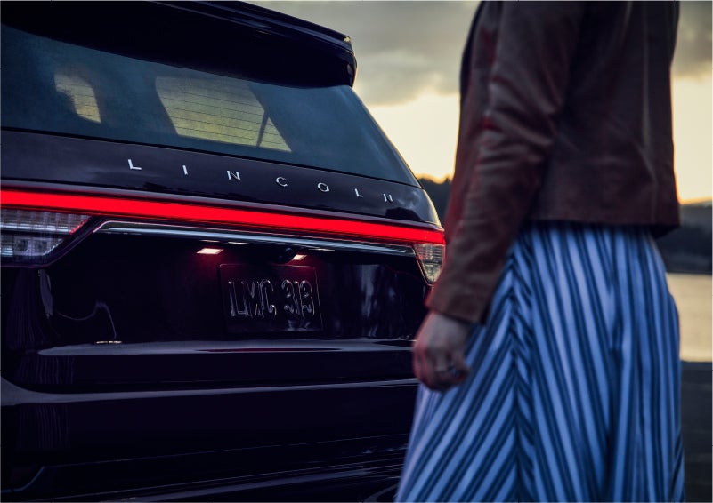 A person is shown near the rear of a 2023 Lincoln Aviator® SUV as the Lincoln Embrace illuminates the rear lights | Cummins Lincoln in Weatherford OK